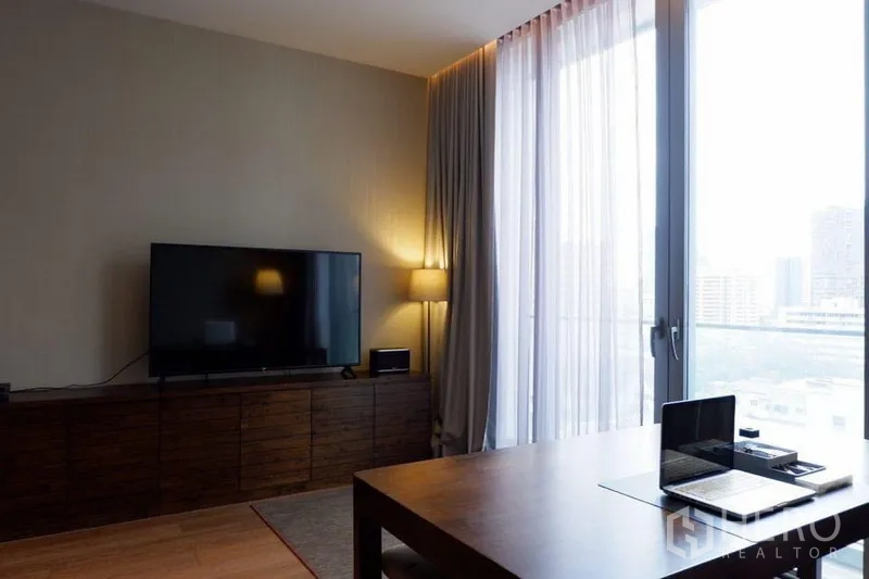 Condominium for sale in Khlong Toei, Bangkok - Living area with long wooden TV console, floor-to-ceiling windows with sheer curtains, and a city view.