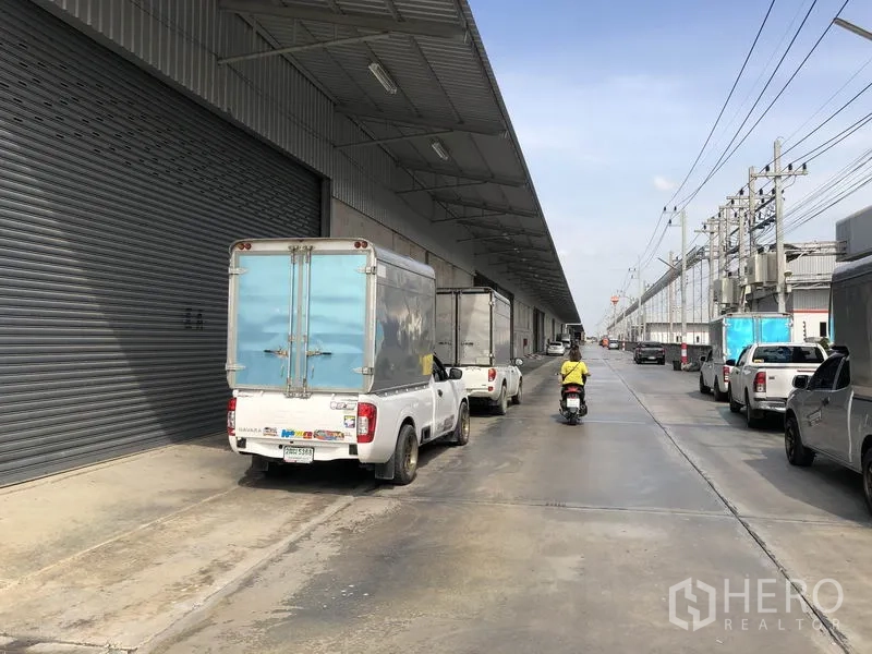 货栈 出租在Lam Luk Ka, Pathum Thani - 仓库外部带遮雨装卸平台和大型卷帘门，配送车辆停靠。