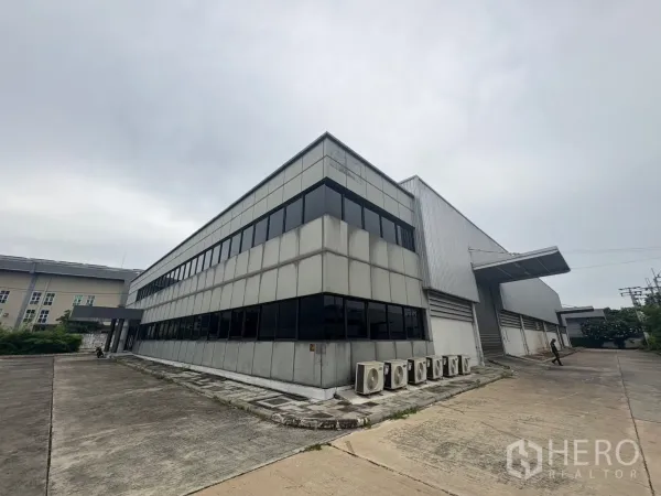 Factory for rent or sale in Samut Prakan - Corner view of the two‑storey office attached to a warehouse with AC condenser units along a concrete driveway.