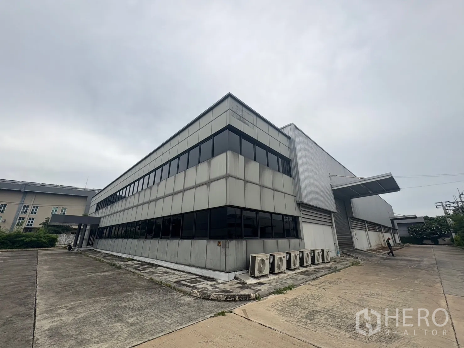 Factory for rent or sale in Samut Prakan - Corner view of the two‑storey office attached to a warehouse with AC condenser units along a concrete driveway.