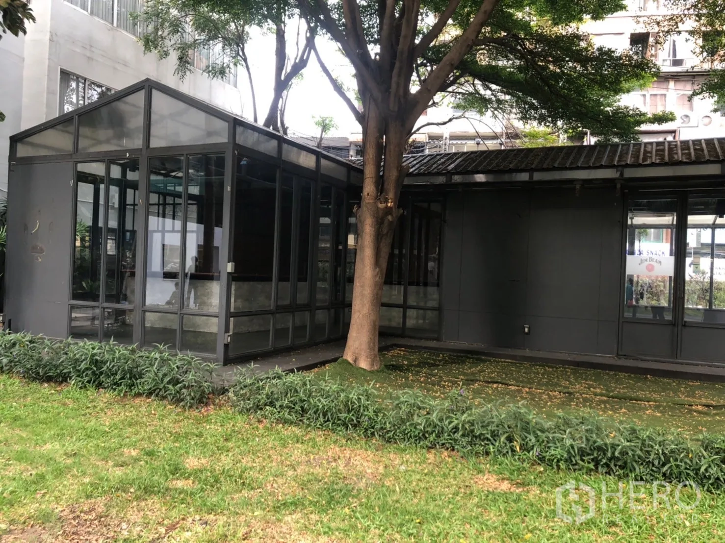 零售空间 出租在Watthana, Bangkok - 玻璃亭与深色建筑外立面，树荫与绿篱点缀的外观。