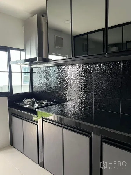 Detached House for rent in Bang Phli, Samut Prakan - Sleek black built-in kitchen with gas hob, hood and textured tile backsplash beside a window.