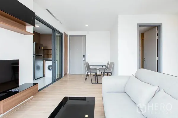 Condominium for rent in Watthana, Bangkok - Living room facing dining set and a separate kitchen behind sliding glass with fridge and washing machine.