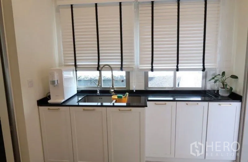 Detached House for rent or sale in Saphan Sung, Bangkok - Secondary kitchen with white cabinets, black worktop, and sink under window blinds.