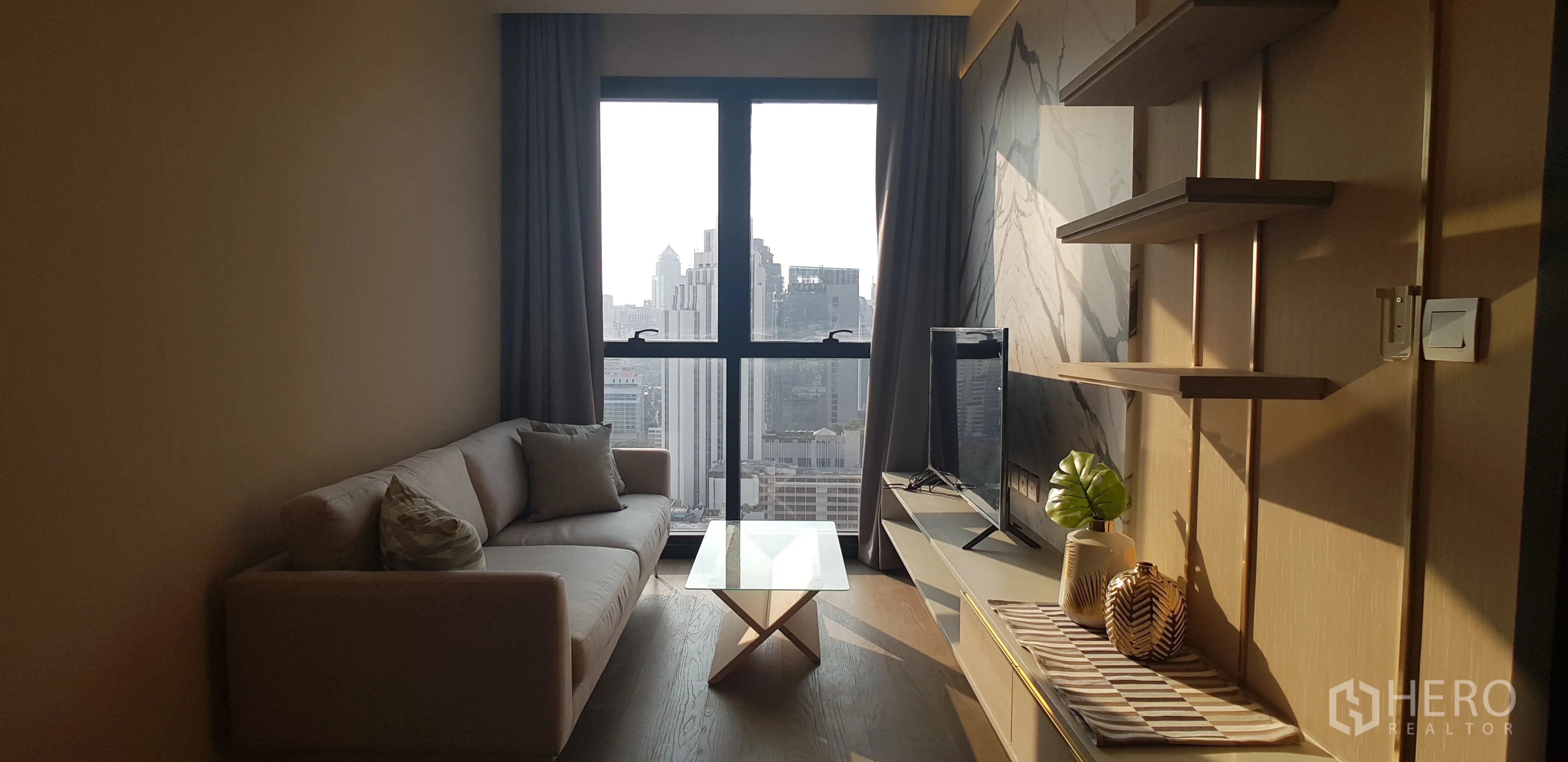 Condominium for rent in Watthana, Bangkok - Close-up of sofa and TV beside a marble accent wall, with a wide city view through the large window.