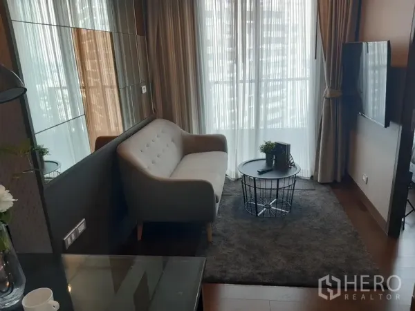 คอนโด ให้เช่าในKhlong Toei, Bangkok - ห้องนั่งเล่นสไตล์โมเดิร์น โซฟาสีเทา โต๊ะกลางทรงลวด พรม ทีวีติดผนัง และหน้าต่างกระจกเต็มบานพร้อมผ้าม่านวิวเมือง