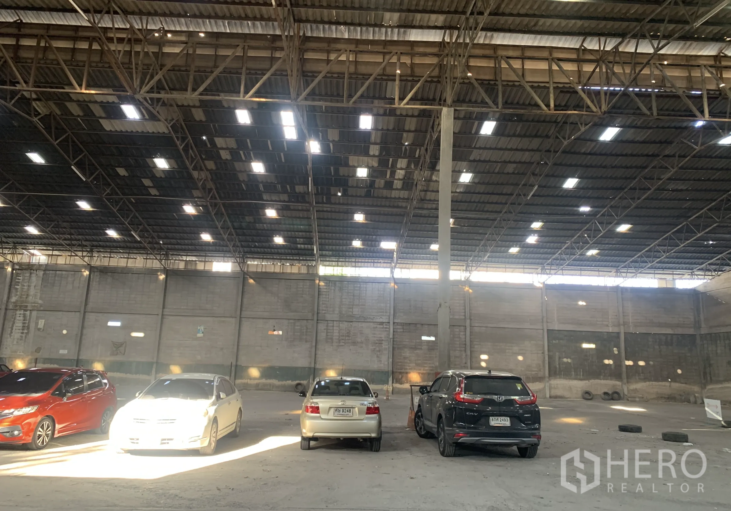 Warehouse for rent in Yan Nawa, Bangkok - Large warehouse hall with high roof, central column, lighting panels, and several cars parked inside.