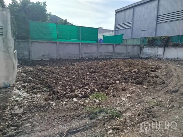 Detached House for rent or sale in Bang Na, Bangkok - Corner of the land plot showing leveled soil and privacy wall beside an industrial-style building.
