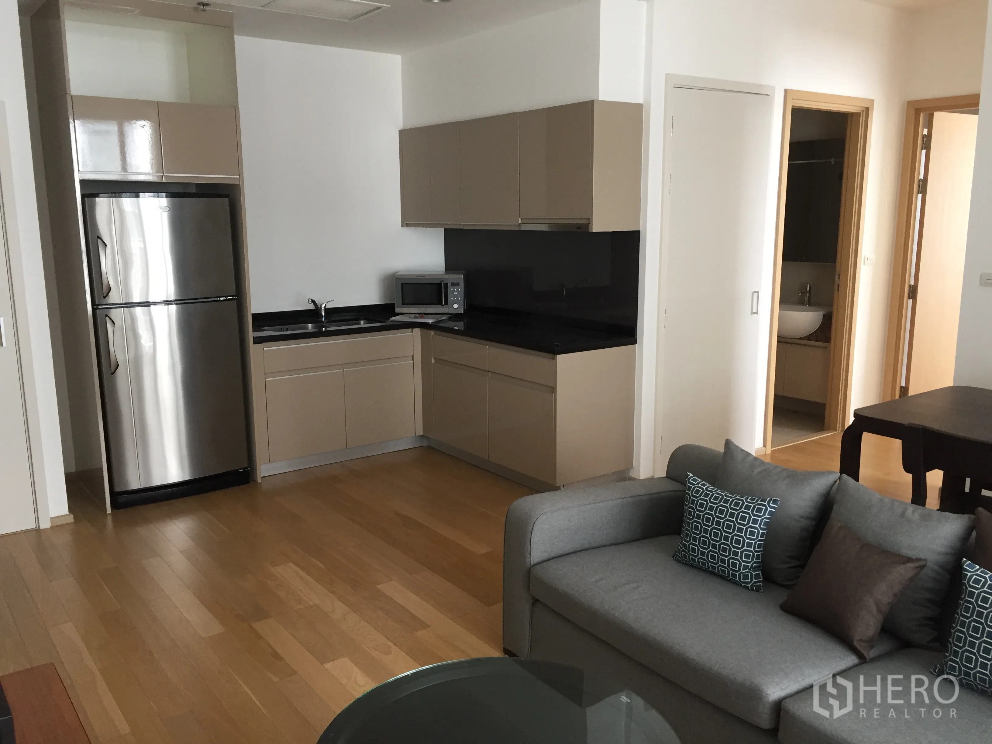 公寓 出租或出售在Watthana, Bangkok - 开放式厨房，米色橱柜与黑色台面，木地板，配不锈钢冰箱与灰色沙发。