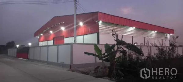 货栈 出租或出售在Khlong Luang, Pathum Thani - 傍晚侧视图，红色金属外墙仓库，围墙内，沿安静道路设有路灯。