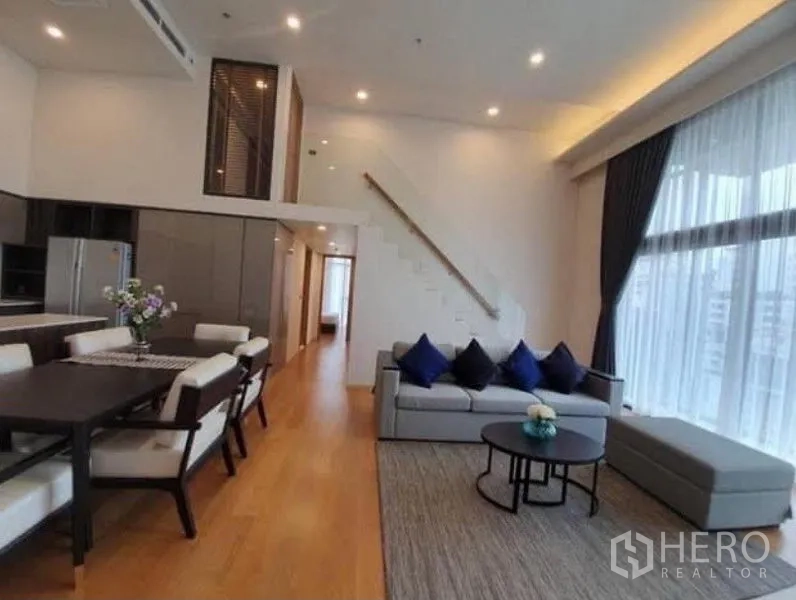 Condominium for rent in Watthana, Bangkok - Spacious lounge and dining area with wood floors, blue accent cushions, and staircase leading to bedrooms.