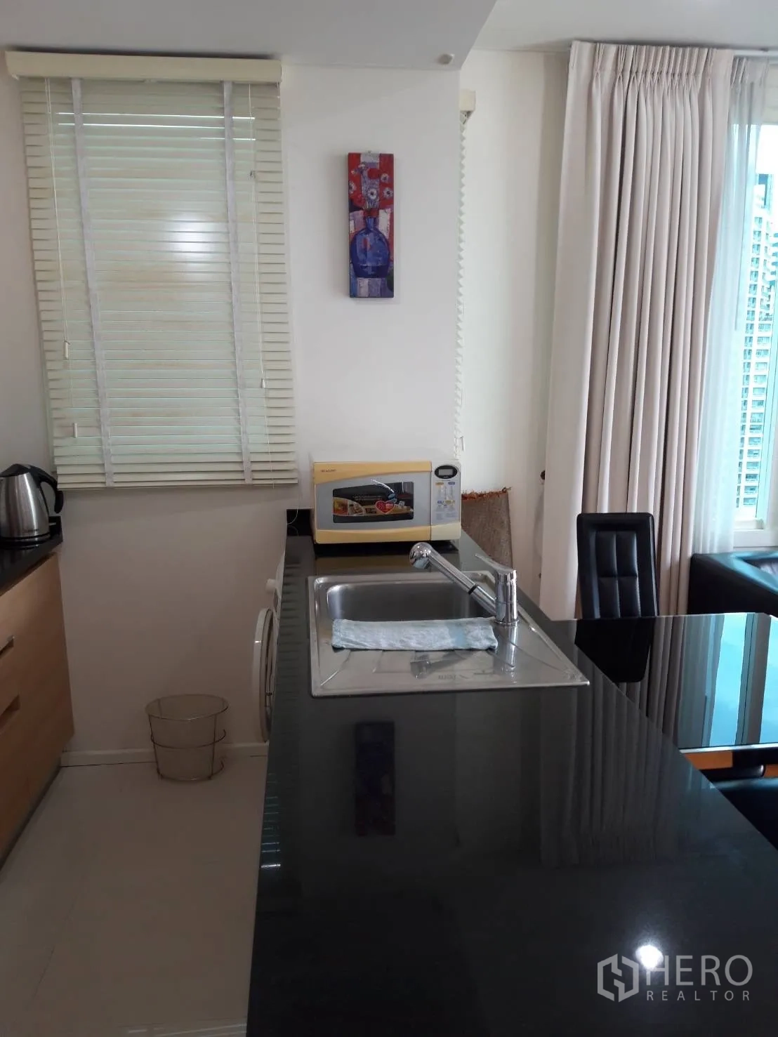 Condominium for rent in Watthana, Bangkok - Long kitchen counter with sink, microwave and window blinds beside living area.