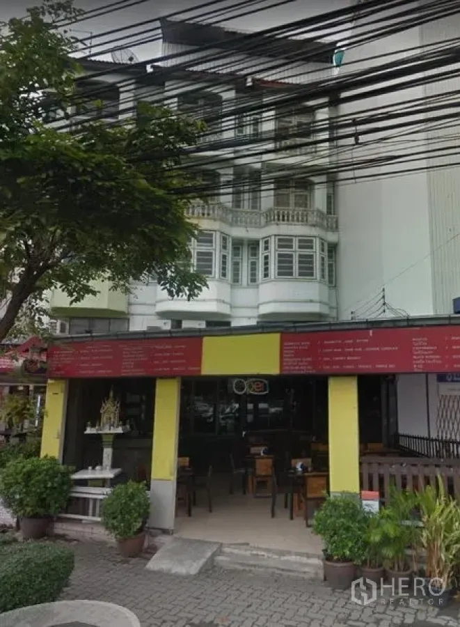 อาคารพาณิชย์ ให้เช่าในKhlong Toei, Bangkok - อาคารพาณิชย์สีขาวหลายชั้น มีหน้าต่างบานโค้ง ชั้นล่างเป็นหน้าร้านเปิดโล่ง มีโต๊ะนั่ง กระถางต้นไม้หน้าร้าน ต้นไม้ริมถนน และสายไฟเหนือศีรษะ.