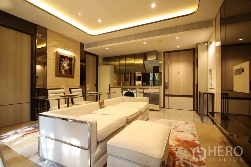 Condominium for rent in Watthana, Bangkok - Open-plan lounge with sofa and ottoman facing a modern kitchen with built-in appliances and warm cove lighting.