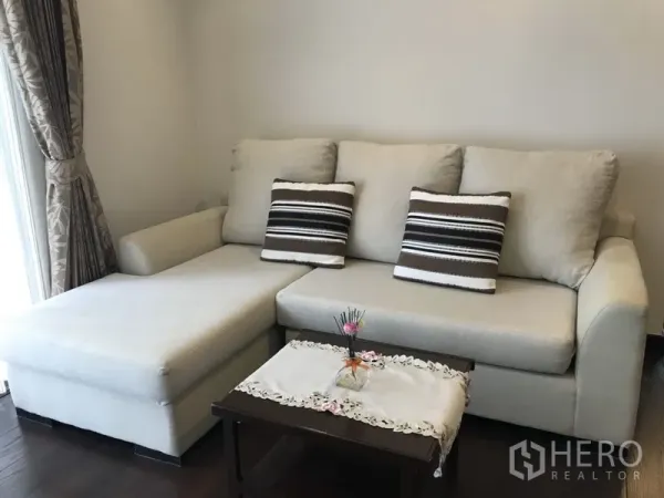 Condominium for rent in Ratchathewi, Bangkok - Beige L-shaped sofa with striped cushions and a coffee table beside a curtained window in the condo living area.