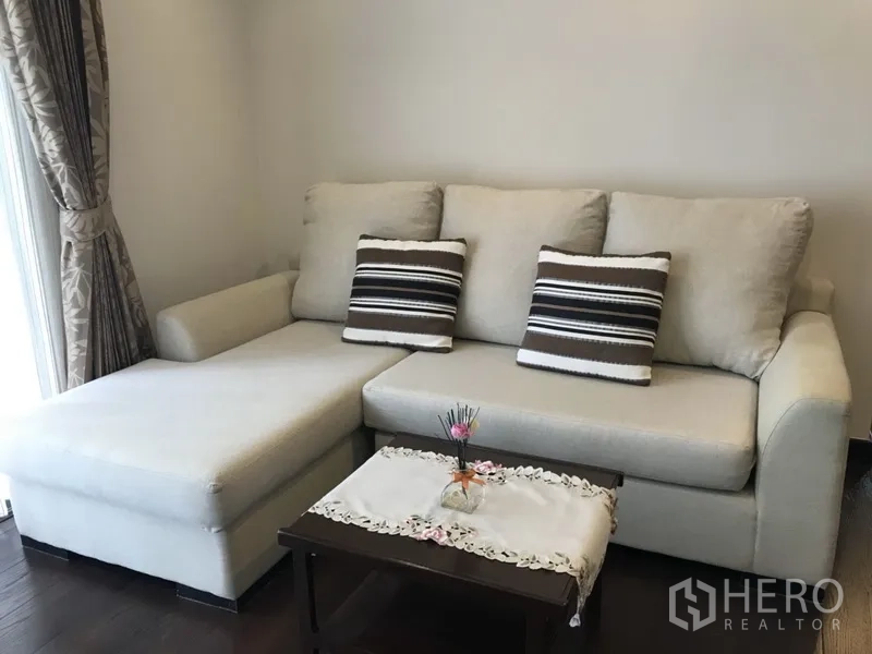 Condominium for rent in Ratchathewi, Bangkok - Beige L-shaped sofa with striped cushions and a coffee table beside a curtained window in the condo living area.