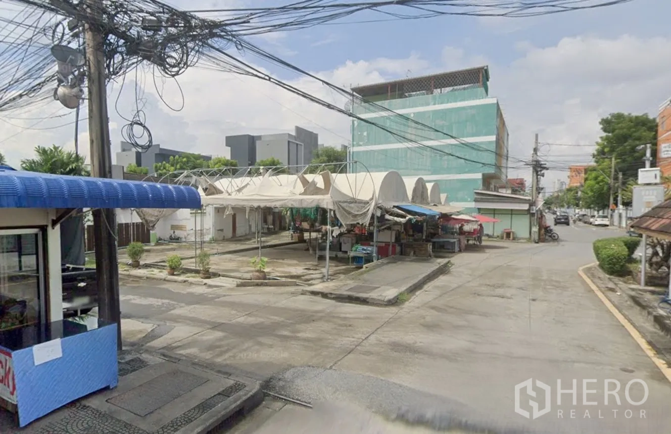 Land for rent in Bang Kapi, Bangkok - Intersection scene showing tented structures on the site, overhead cables, and small shops.