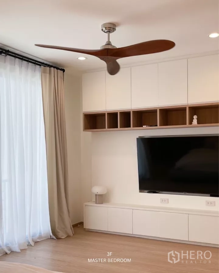 ทาวน์เฮาส์ ให้เช่าในSuan Luang, Bangkok - ผนังทีวีห้องนอนมาสเตอร์พร้อมชั้นบิวท์อิน ทีวีจอใหญ่ และพัดลมเพดานใกล้ผ้าม่านโปร่ง