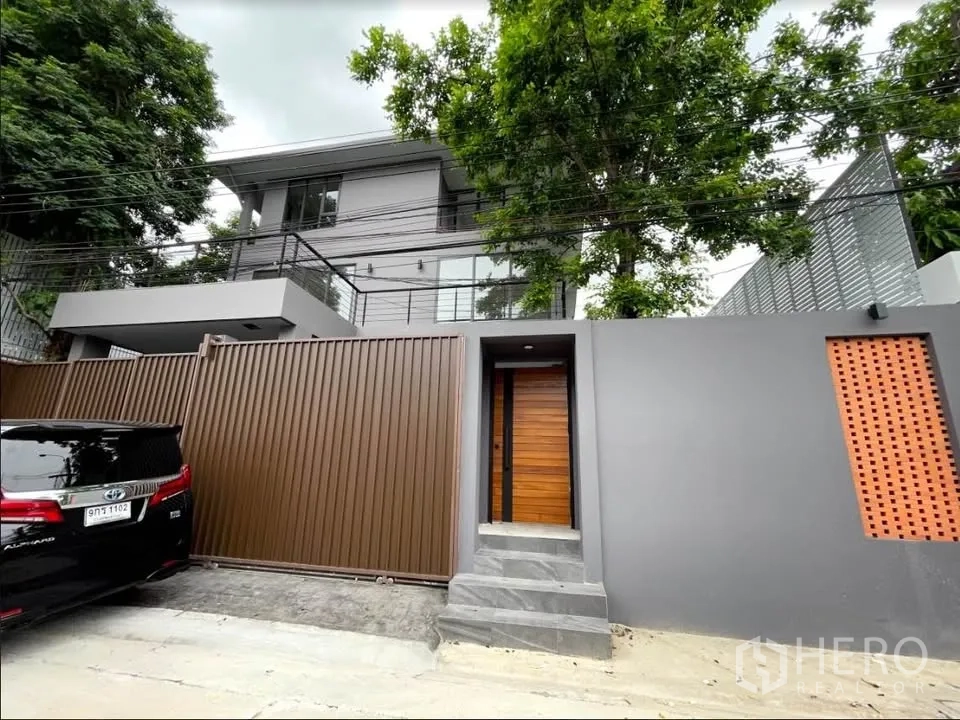 Detached House for sale in Watthana, Bangkok - Modern gated facade of a multi-level detached house with tall privacy wall and trees.