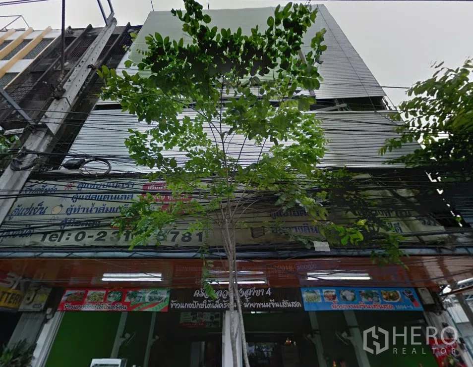 อาคารพาณิชย์ ให้เช่าในPhra Khanong, Bangkok - ด้านหน้าระดับถนนของอาคารพาณิชย์พร้อมประตูม้วนและป้ายร้าน ใต้ร่มไม้