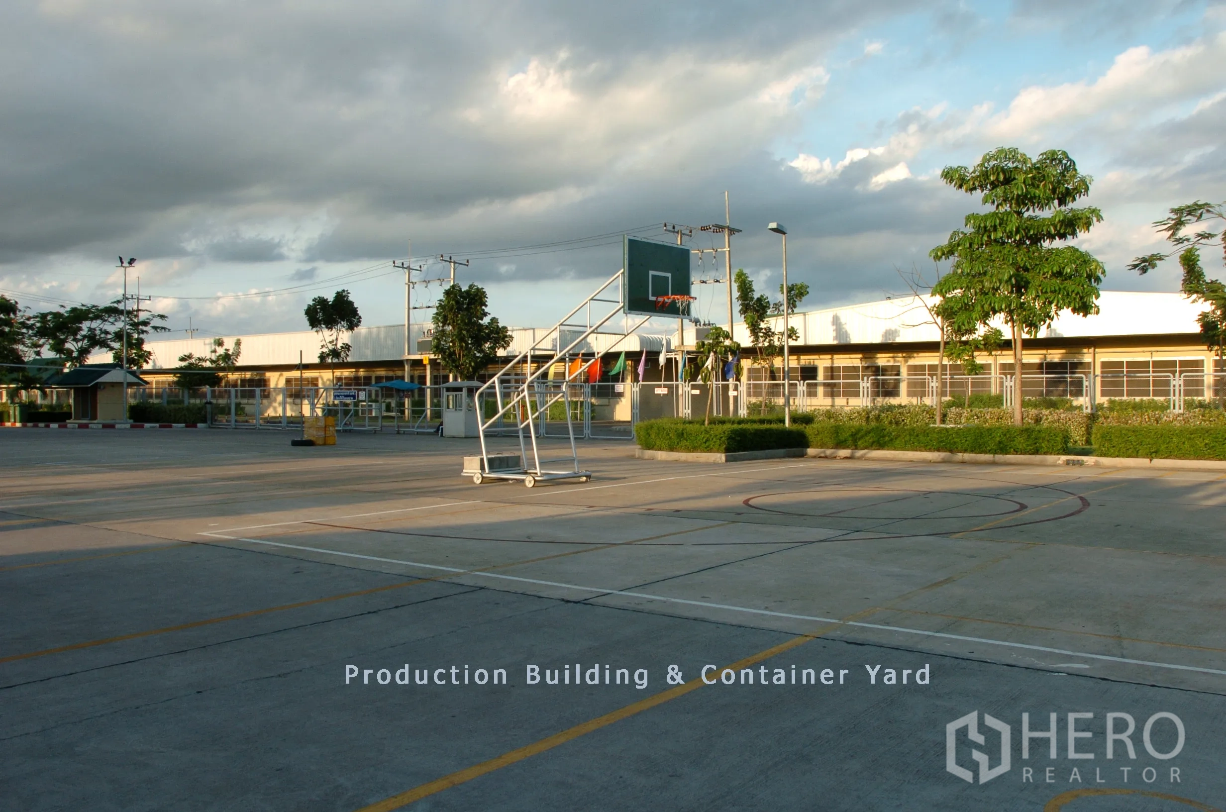 工厂 for sale in Khao Chamao, Rayong - 宽阔的集装箱作业场与生产大楼，带可移动篮球架、围栏与绿化，天空多云。