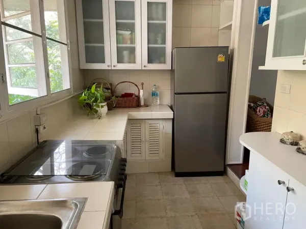 公寓 出租在Watthana, Bangkok - 明亮的封闭式厨房，带大窗户、玻璃门吊柜、灶台和不锈钢冰箱。