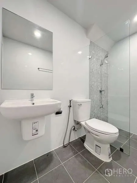 Townhouse for rent in Min Buri, Bangkok - Modern bathroom with glass-enclosed shower, white basin and toilet on gray tile flooring.