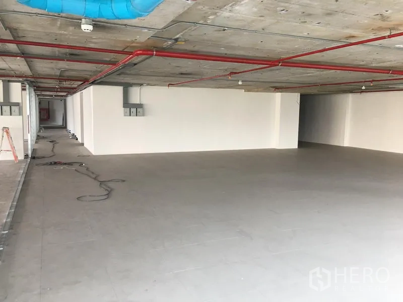 พื้นที่ขายปลีก ให้เช่าในKhlong Toei, Bangkok - พื้นที่ค้าปลีกแบบโอเพ่นสเปซ พื้นกระเบื้องสีเทา ผนังสีขาว เพดานคอนกรีตเปลือยพร้อมท่อสprinkler สีแดงและท่อดักท์สีน้ำเงิน.