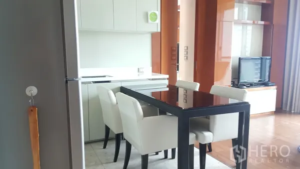 公寓 出租或出售在Khlong Toei, Bangkok - 开放式厨房与餐区，白色橱柜、黑色玻璃餐桌和四把软包椅，连接带电视的起居区。