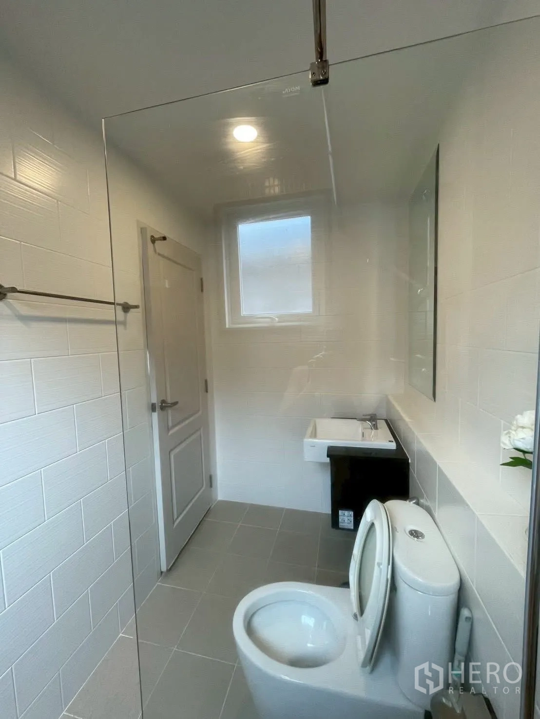 Detached House for rent in Prawet, Bangkok - Narrow bathroom with toilet, sink and glass shower partition beneath a window.