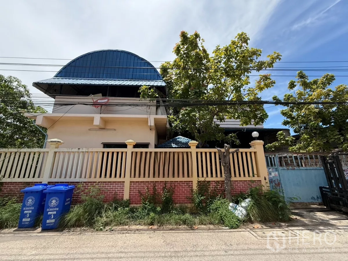 Warehouse for sale in Phra Samut Chedi, Samut Prakan - Street view of gated compound with tall fence, mature trees and a blue arched warehouse roof.