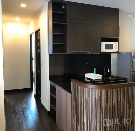 Condominium for rent in Ratchathewi, Bangkok - Modern kitchenette with dark wood cabinets, black countertop, microwave and a curved breakfast bar.