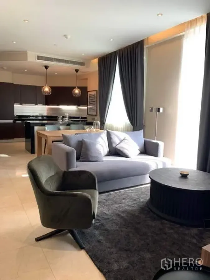 Condominium for rent in Bang Rak, Bangkok - Open-plan living area with gray sofa, round coffee table, floor-to-ceiling curtains, and view to a modern kitchen and dining zone.
