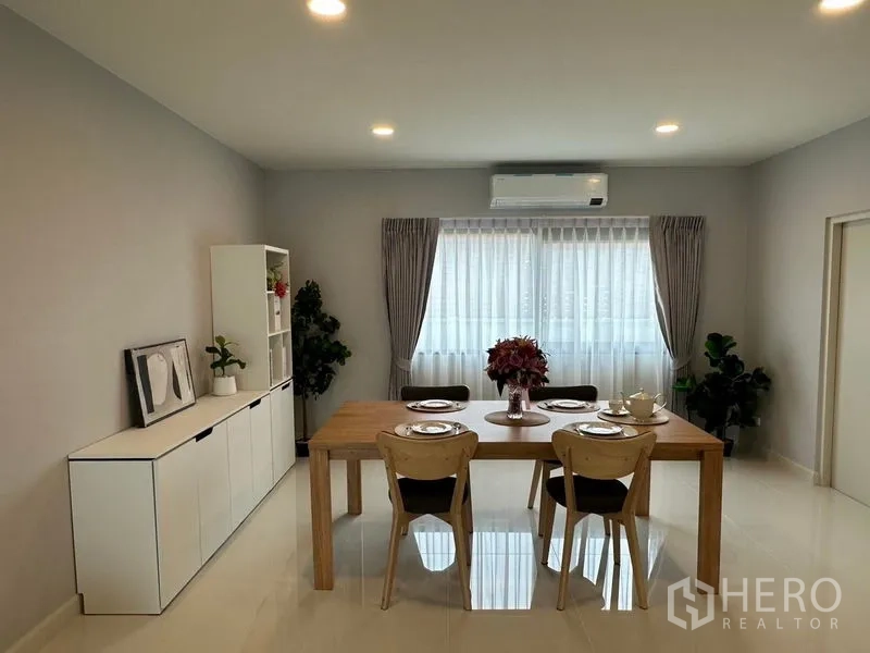 Detached House for rent in Bang Phli, Samut Prakan - Dining room with wooden table set for four, white sideboard and a large window dressed with sheer curtains.
