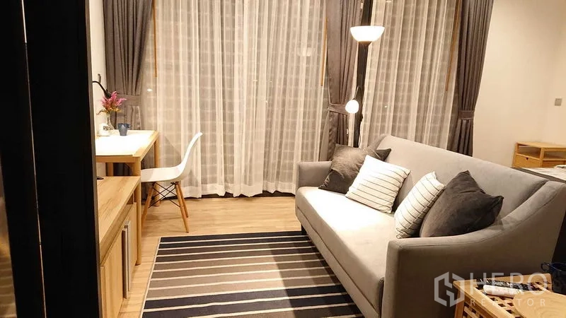 คอนโด ขายในWatthana, Bangkok - มุมนั่งเล่นอบอุ่น โซฟาสีเทา พรมลายทาง ผ้าม่านสูง และมุมทำงานข้างหน้าต่าง