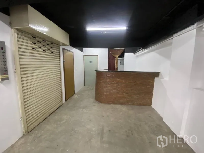 零售空间 出租在Bangkok - 店铺室内带接待台与卷闸门，白色墙面与混凝土地面，日光灯照明。