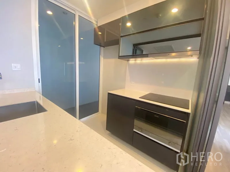 公寓 出租在Bang Rak, Bangkok - 紧凑厨房区，亮面柜体、电磁炉与内置烤箱，灯带照明。