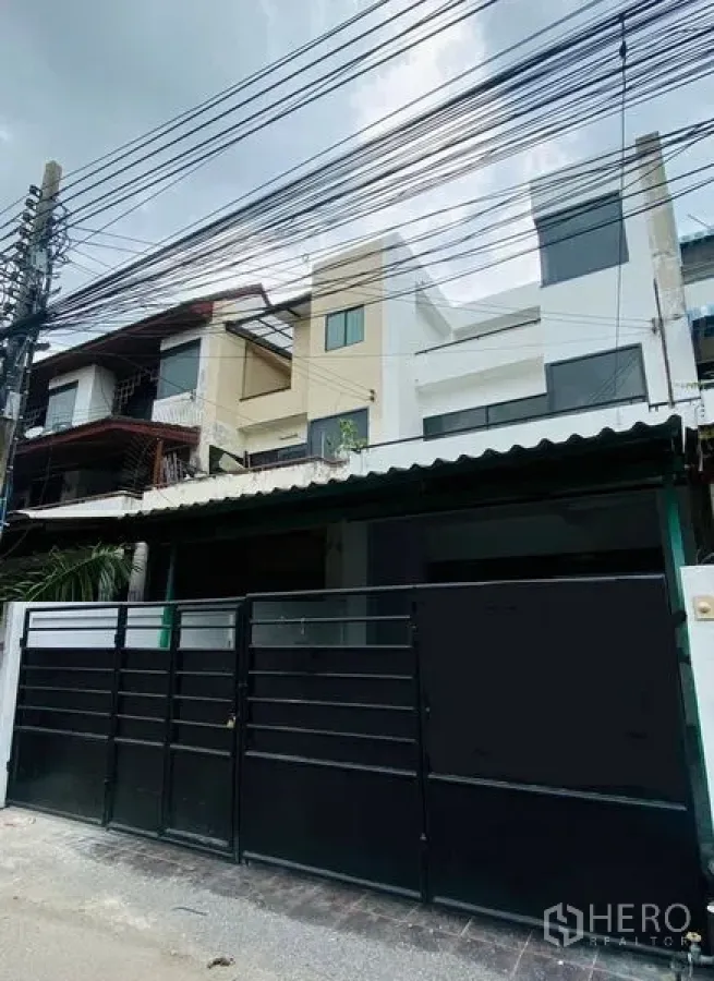 สำนักงานที่บ้าน ให้เช่าหรือขายในSuan Luang, Bangkok - มุมมองหน้าบ้านสามชั้น พร้อมรั้วเหล็กสีดำและสายไฟเหนือศีรษะ