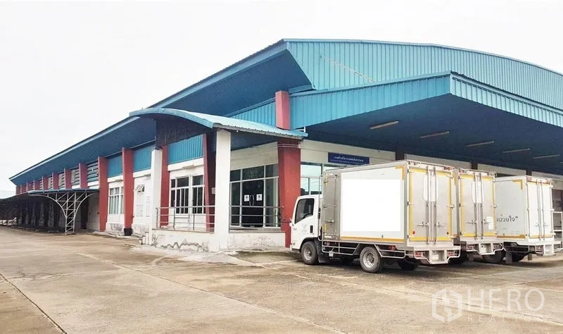 โรงงาน ขายในLam Luk Ka, Pathum Thani - อาคารโรงงานหลังคาเมทัลชีทสีน้ำเงิน ด้านหน้าเป็นสำนักงานกระจกและท่าขนถ่ายสินค้าแบบยกพื้น มีรถบรรทุกจอด