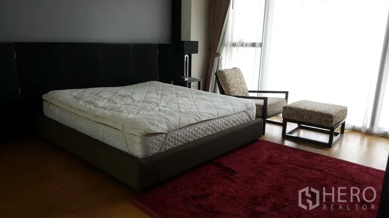 Condominium for rent in Sathon, Bangkok - Master bedroom with king bed, accent chair, red rug, and full-height windows for abundant light.