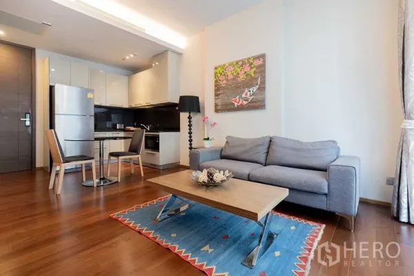 Condominium for rent - Bright open-plan living room with gray sofa, wood floors, colorful rug, and fitted kitchen in a Thonglor condo.