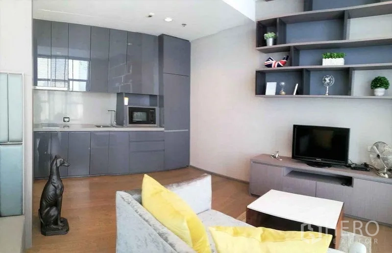 Condominium for sale in Bang Rak, Bangkok - Open-plan living area with sleek gray kitchen cabinets, wall shelves, TV console, and sofa with yellow cushions.