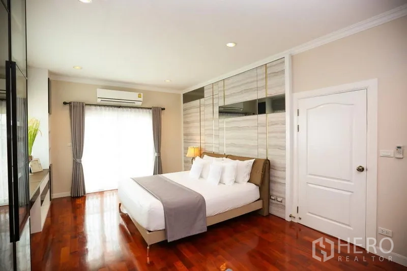 Detached House for rent in Bang Na, Bangkok - Master bedroom with feature wall, hardwood floors, king bed and curtains over a balcony window.