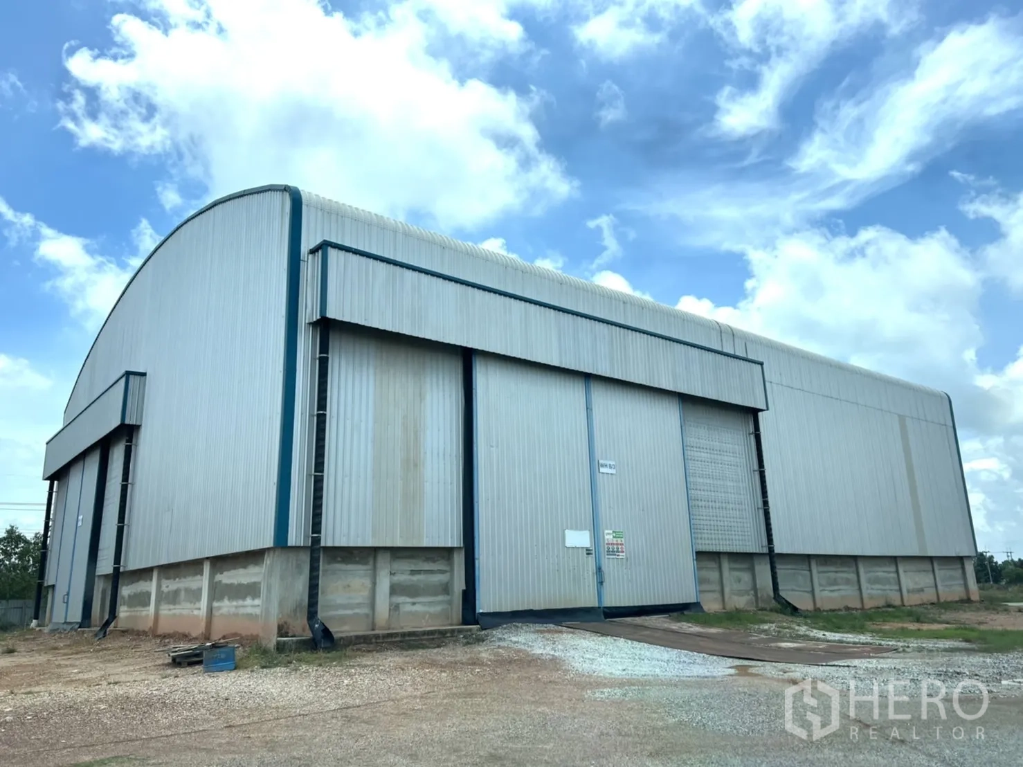 Warehouse for rent in Ban Pho, Chachoengsao - Steel-clad warehouse exterior with high sliding doors and a gravel yard under a bright sky.