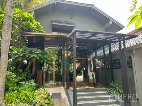 零售空间 出租在Bangkok - 现代玻璃门廊，绿植环绕，旁为坡顶建筑。