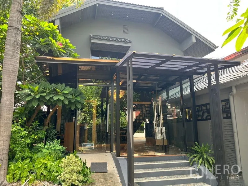 零售空间 出租在Bangkok - 现代玻璃门廊，绿植环绕，旁为坡顶建筑。