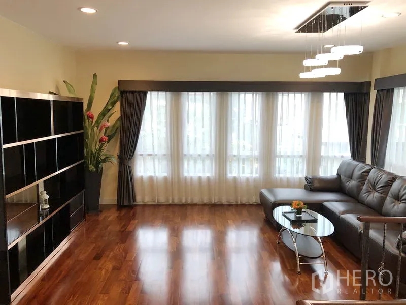 Detached House for rent or sale in Saphan Sung, Bangkok - Upstairs family room with wood floors, L-shaped sofa, large windows and shelving unit.