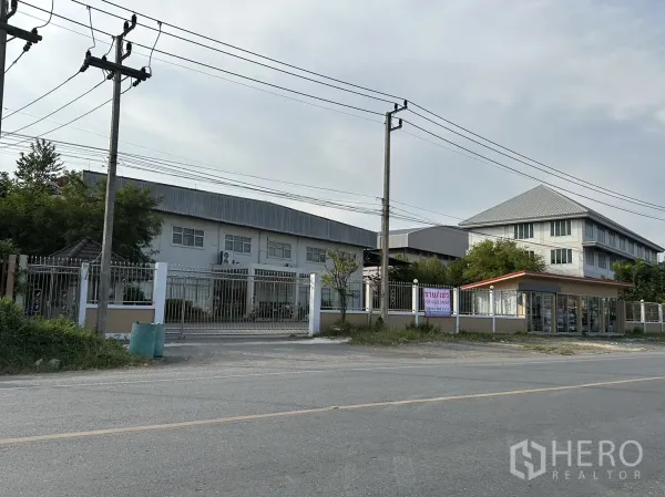 โรงงาน ให้เช่าหรือขายในLam Luk Ka, Pathum Thani - ด้านหน้าโรงงานมุมเอียง เห็นประตูรั้ว รั้วรอบพื้นที่ และป้อมยามข้างอาคารหลัก