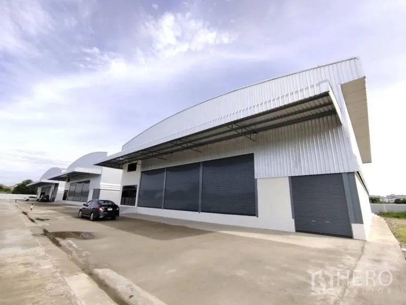 Warehouse for rent in Bang Phli, Samut Prakan - Modern warehouse exterior with curved metal roof, wide driveway and multiple roller shutter doors.