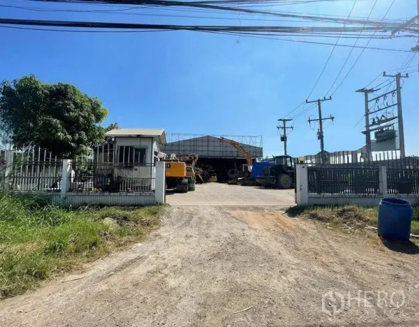 货栈 出租在Wang Noi, Phra Nakhon Si Ayutthaya - 仓库大门与警卫室，宽阔车道通往钢结构棚厂，院内停放重型机械，电线杆沿路，蓝天晴朗。